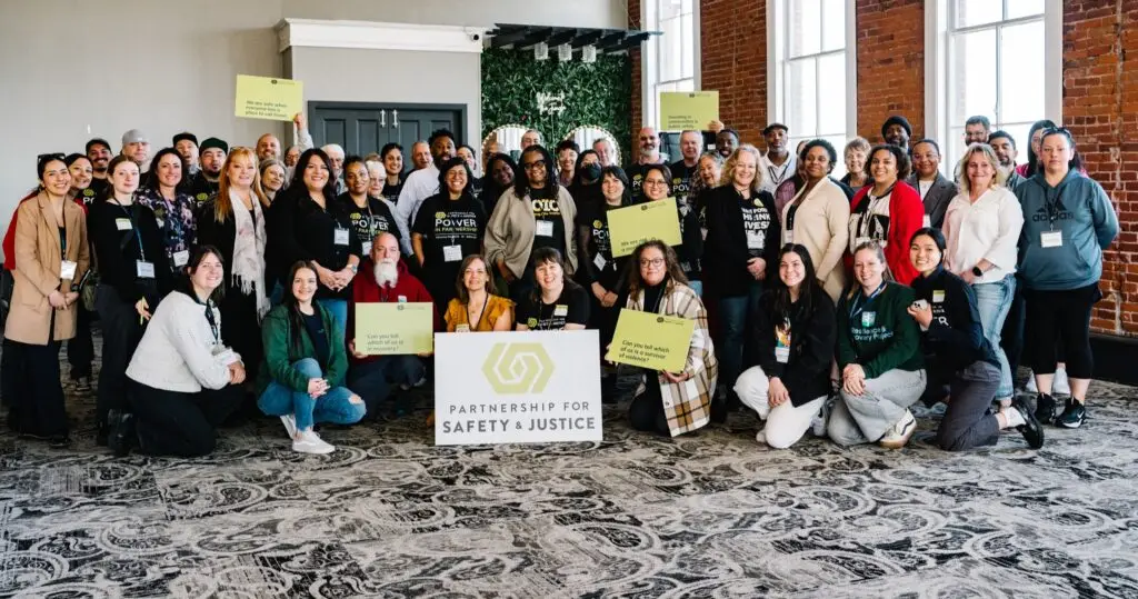 Indoor group picture of more than 50 people with a brick wall with bright windows and foliage in the background. Some people are holding green signs with unreadable text.